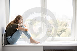 Sad depressed young woman having social problems sitting on windowsill