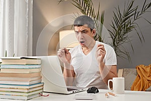 Sad despair Caucasian adult man wearing white T-shirt working at home with laptop making mistake screaming no with negative