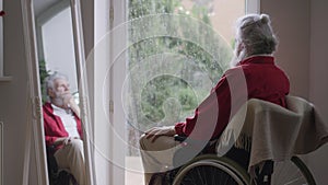 Sad disabled bearded grey-haired old man looking at rain sitting in wheelchair indoors. Frustrated upset Caucasian