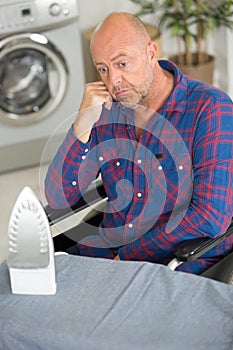 Sad disabled man sitting in wheelchair ironing shirt