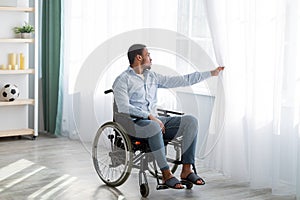 Sad disabled young man sitting on wheelchair alone at home, looking through window, full length portrait
