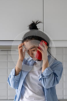 Sad disappointed Asian girl listening bad news over phone while standing in kitchen at home