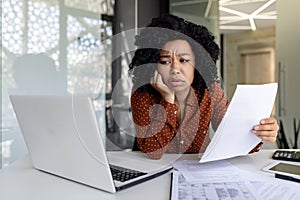 Sad and disappointed businesswoman inside office at workplace looking at documents papers financial reports, african