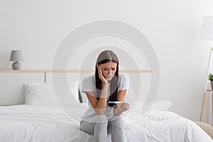 Sad disappointment young european woman hold pregnancy test, sits on bed alone in bedroom