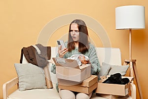 Sad displeased woman sitting on sofa among boxes with clothing against beige wall using mobile phone checking her order with