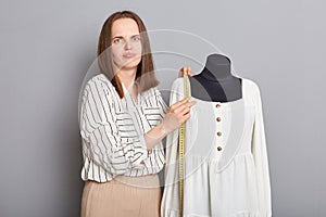 Sad dissatisfied young adult female sewer standing near mannequin with new dress isolated over gray background, looking at camera