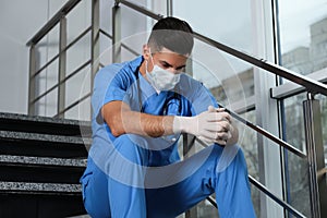 Sad doctor in facial mask on stairs indoors. Stress of health care workers during coronavirus pandemic