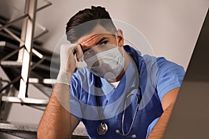 Sad doctor in facial mask on stairs indoors. Stress of health care workers during coronavirus pandemic