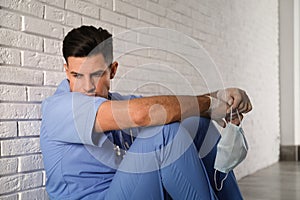 Sad doctor with protective mask sitting near white brick wall indoors. Stress of health care workers during coronavirus pandemic