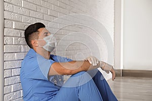 Sad doctor sitting near white brick wall indoors. Stress of health care workers during coronavirus pandemic