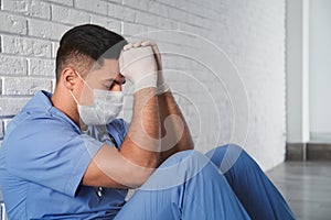 Sad doctor sitting near white brick wall indoors. Stress of health care workers during coronavirus pandemic