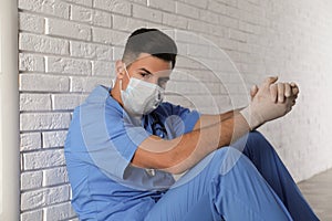 Sad doctor sitting near white brick wall indoors. Stress of health care workers during coronavirus pandemic