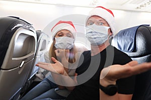Sad and drunk man and woman in protective medical masks and santa claus red caps flying in airplane