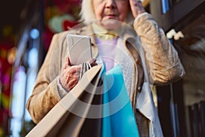 Sad elderly female standing on the street while waiting call on her smartphone