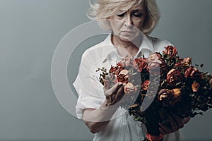 Sad elderly woman touch bouquet of withered roses