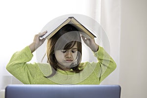 Sad elementary schooler girl studying at home with a book on her head because she is tired and angry - Little girl studying online