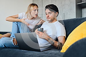 Sad emotional blond woman angrily talking to calm brunette man with cup of coffee in hands and laptop near. Young couple