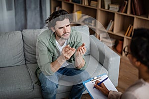 Sad emotional young european man gesturing and talking to black female psychologist in clinic interior