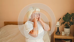 Sad european 60s senior female, worried, suffers from insomnia and headache. Lady sits on bed in bedroom at home