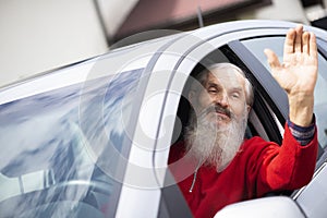 Sad face bearded senior man waving hand through window of gray car