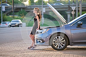 Sad female driver standing on a city street near her car with popped up hood and broken engine waiting for road service