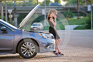 Sad female driver standing on a city street near her car with popped up hood and broken engine waiting for road service