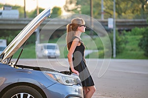 Sad female driver standing on a city street near her car with popped up hood and broken engine waiting for road service