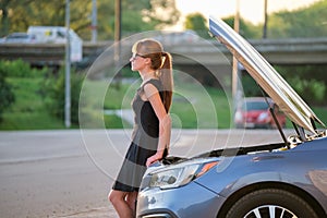 Sad female driver standing on a city street near her car with popped up hood and broken engine waiting for road service