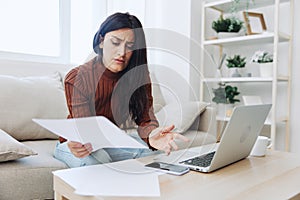 A sad freelance woman with a laptop on her desk works online with documents and invoices from home shrugs her hands in