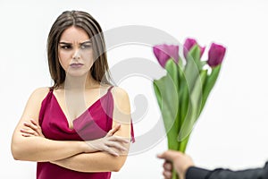 Sad frowning young woman standing with arms crossed and looks displeaased at the tulips man gives her over white