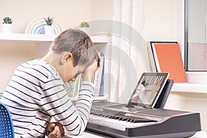 Sad frustrated boy has difficulty learning to play piano at home. Kid using digital tablet. Online learning difficulties photo