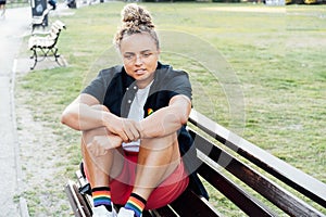 Sad frustrated lesbian woman with LGBTQ rainbow badge on t-shirt sitting on the park bench alone. Concept of loneliness