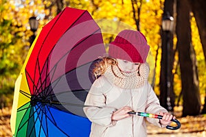 Sad funny beautiful blonde child with rainbow colorful umbrella in a gray jacket in the autumn park. Girl kid playing on