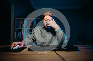 Sad gamer sits at a table at home in a headset on his head and covers his face with his hand from losing the game. Upset boy lost