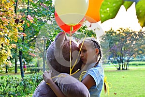 Sad girl child hugs a big teddy bear and balloons in the park. Schoolgirl unhappy in the park with a big toy and balls