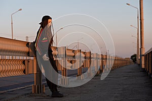 Sad girl with dreadlocks in black sportswear stands on the road at sunset. girl in a cap hitchhiking.