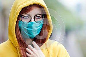 A sad girl with glasses in a reusable medical blue mask looks away warily