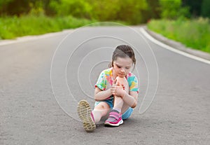 Sad girl hurt her knee and is sitting on the road. child in shorts, a t-shirt and sneakers. bruise on the leg