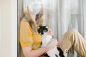 Sad girl in medical protective mask and yellow t-shirt sitting on the windowsill with a black and white Tuxedo cat