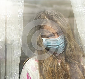 Sad  girl in protective medical mask looks out the window.Sick girl look at the window with longing on the street . Sadness child