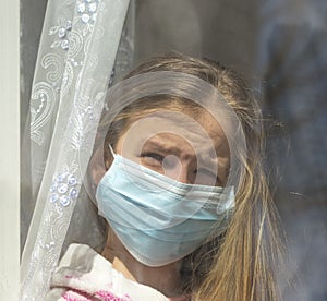 Sad  girl in protective medical mask looks out the window.Sick girl look at the window with longing on the street . Sadness child