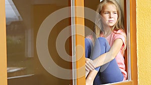 Sad girl sitting at home on the windowsill. Teenage girl waves `hello` through the window