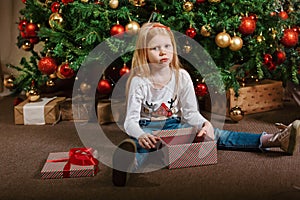 A sad girl is sitting next to a Christmas tree who received an unwanted gift on Christmas Day