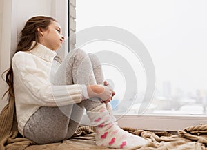 Sad girl sitting on sill at home window in winter