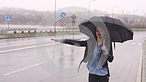 A sad girl is standing under an umbrella on an empty road, she catches the car.