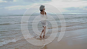 Sad girl walking ocean beach in white bikini on gloomy day. Thinking curly lady