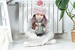 Sad girl in woolen hat, socks and pullover hold hot drink and sit with crossed legs near heating convector, calorifer