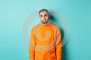 Sad and gloomy man sulking, looking bored at upper left corner, standing in orange sweater against light blue background