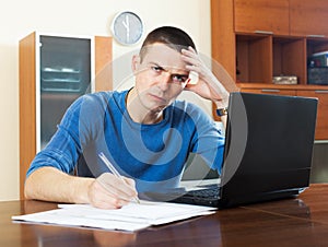 Sad guy with laptop and financial documents at home interior