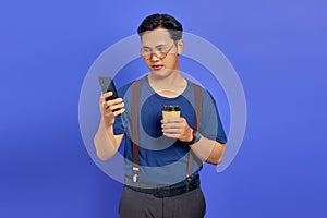 Sad handsome young man holding smartphone and cup of coffee on purple background